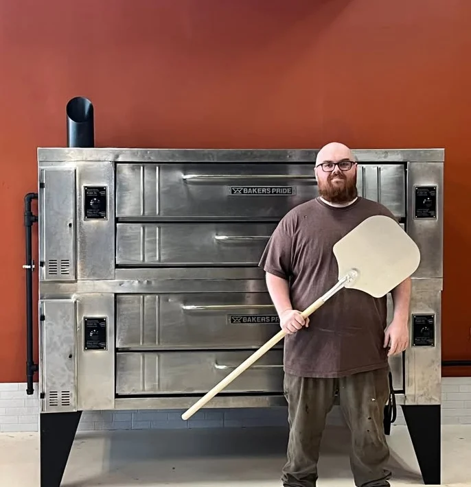 Picture of the owner posing with a large pizza peel in front of double pizza ovens, brand Baker's Pride