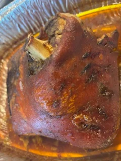 A roasted pork knuckle with crispy skin, resting in a tray with sauce.