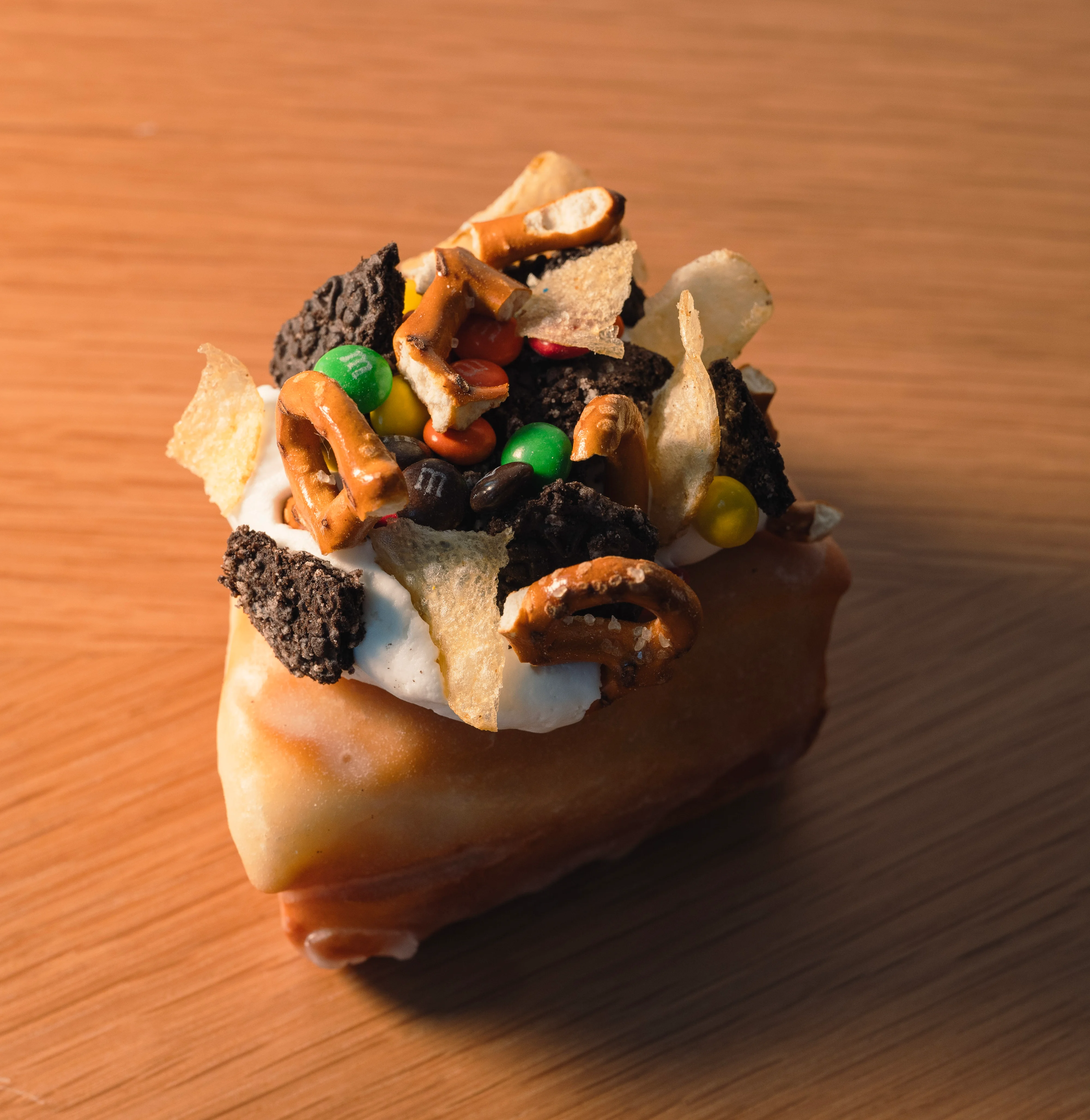 Creative donut topped with pretzels, M&M's, crushed cookies, and potato chips from BAD Bakery, displayed on a wooden surface.