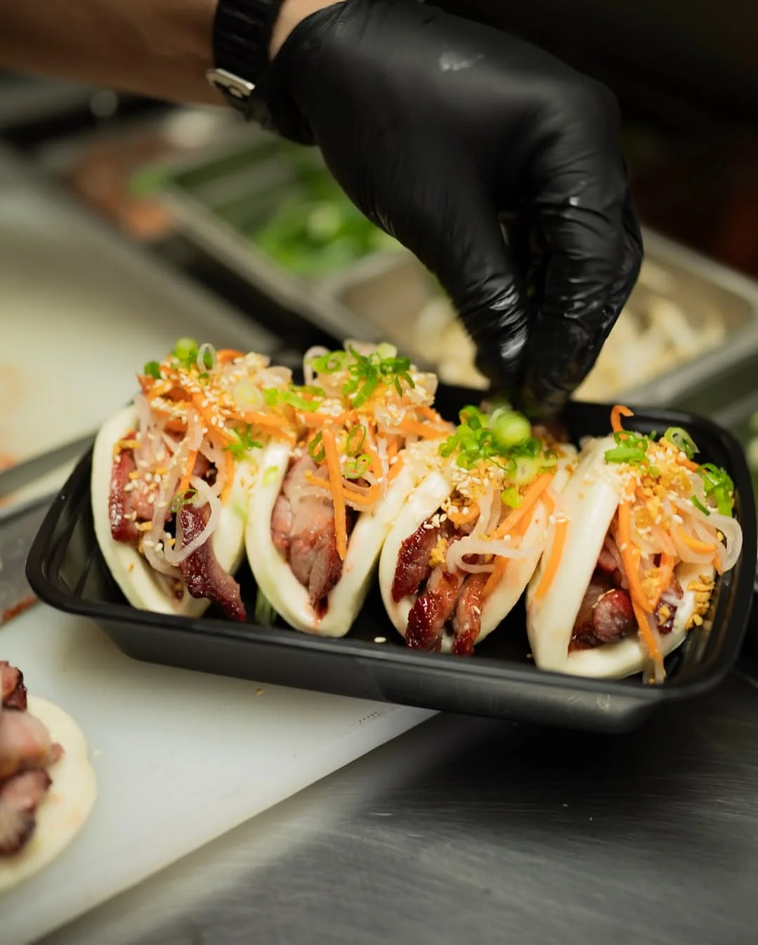 A serving of bao buns filled with meat and topped with fresh vegetables and sesame seeds, presented in a black container.
