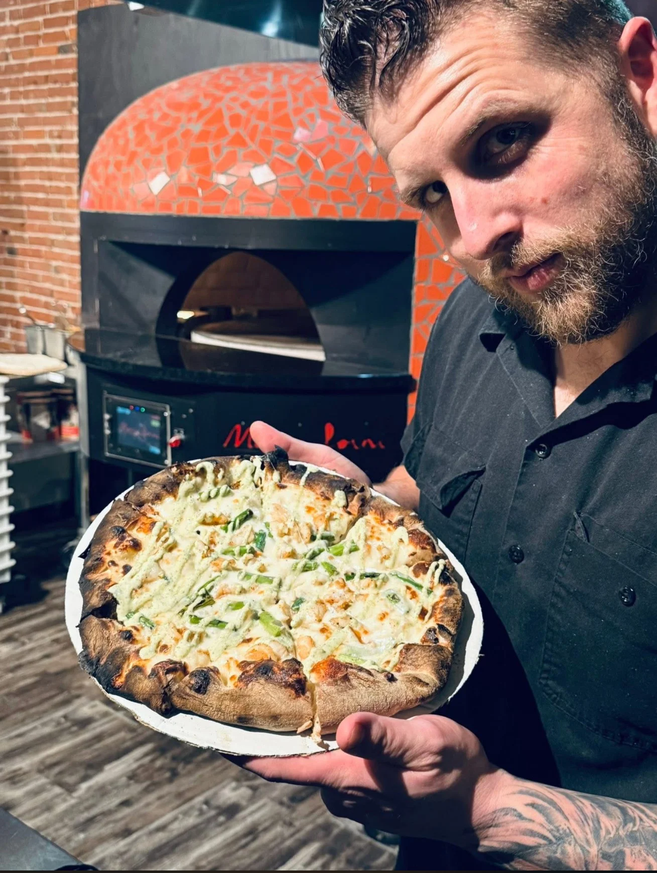 A chef holding a freshly baked pizza with a golden crust and green toppings in front of a pizza oven.