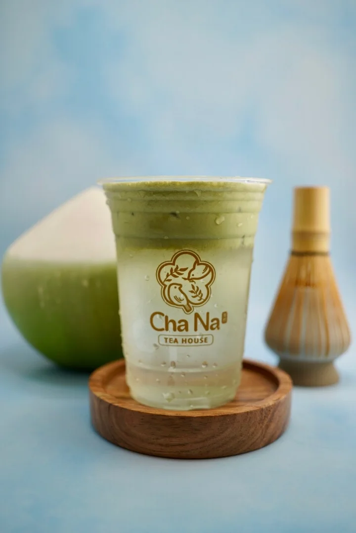 A chilled green tea drink served in a clear cup, accompanied by a bamboo whisk and a whole coconut in the background.