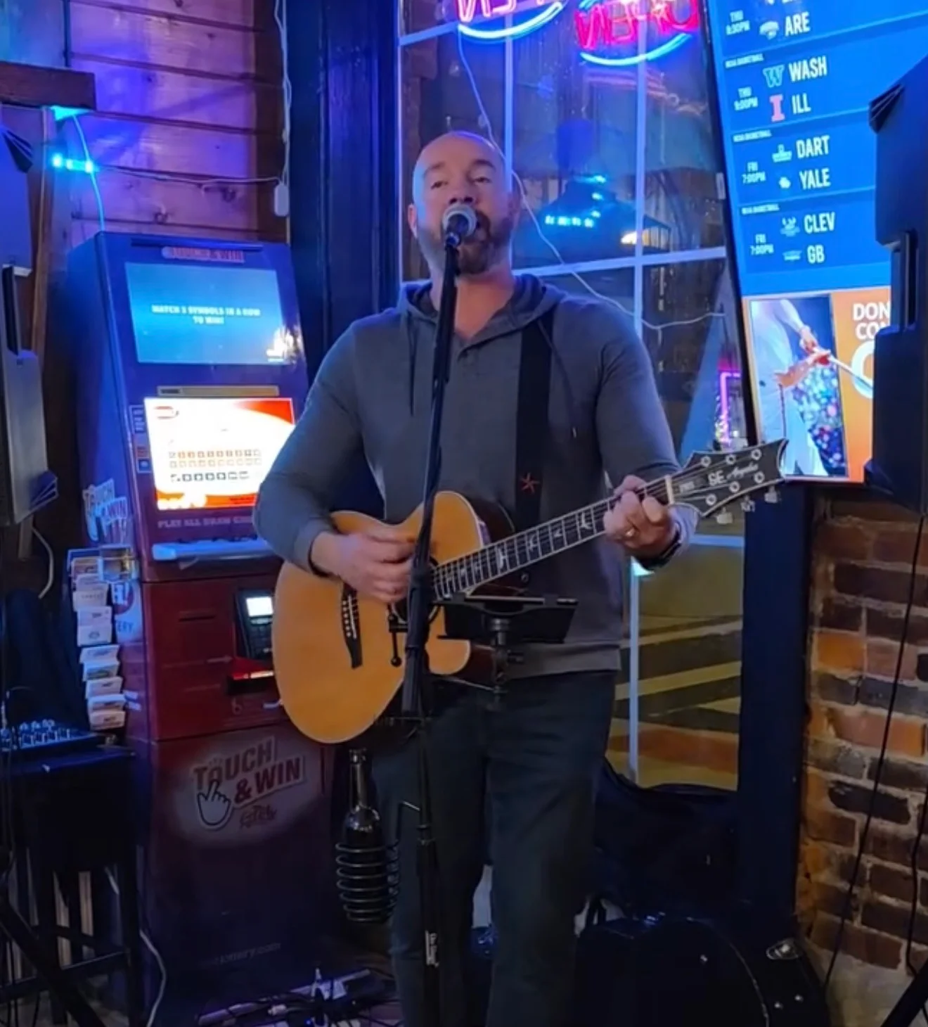 A performer singing into a microphone while playing an acoustic guitar at a lively venue, with neon lights in the background.