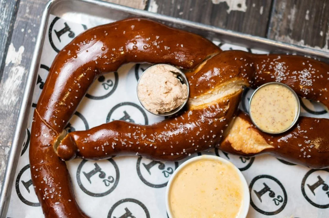 A large soft pretzel served with two dipping sauces, placed on a tray with a branded paper liner.