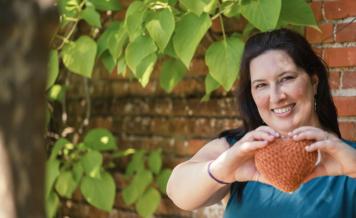 A woman in a teal shirt holds a stuffed crochet heart in her hands. Her fingers are curled to make a heart shape. She stands in front of a brick wall with ivy.