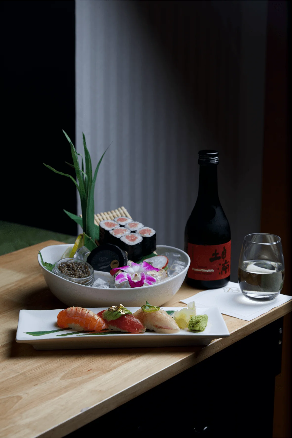 An elegant sushi arrangement featuring three pieces of premium nigiri garnished with gold leaf and microgreens on a white rectangular plate, accompanied by a bowl of maki rolls, caviar, and an orchid blossom over ice, served next to a bottle of sake and a clear glass on a wooden table.