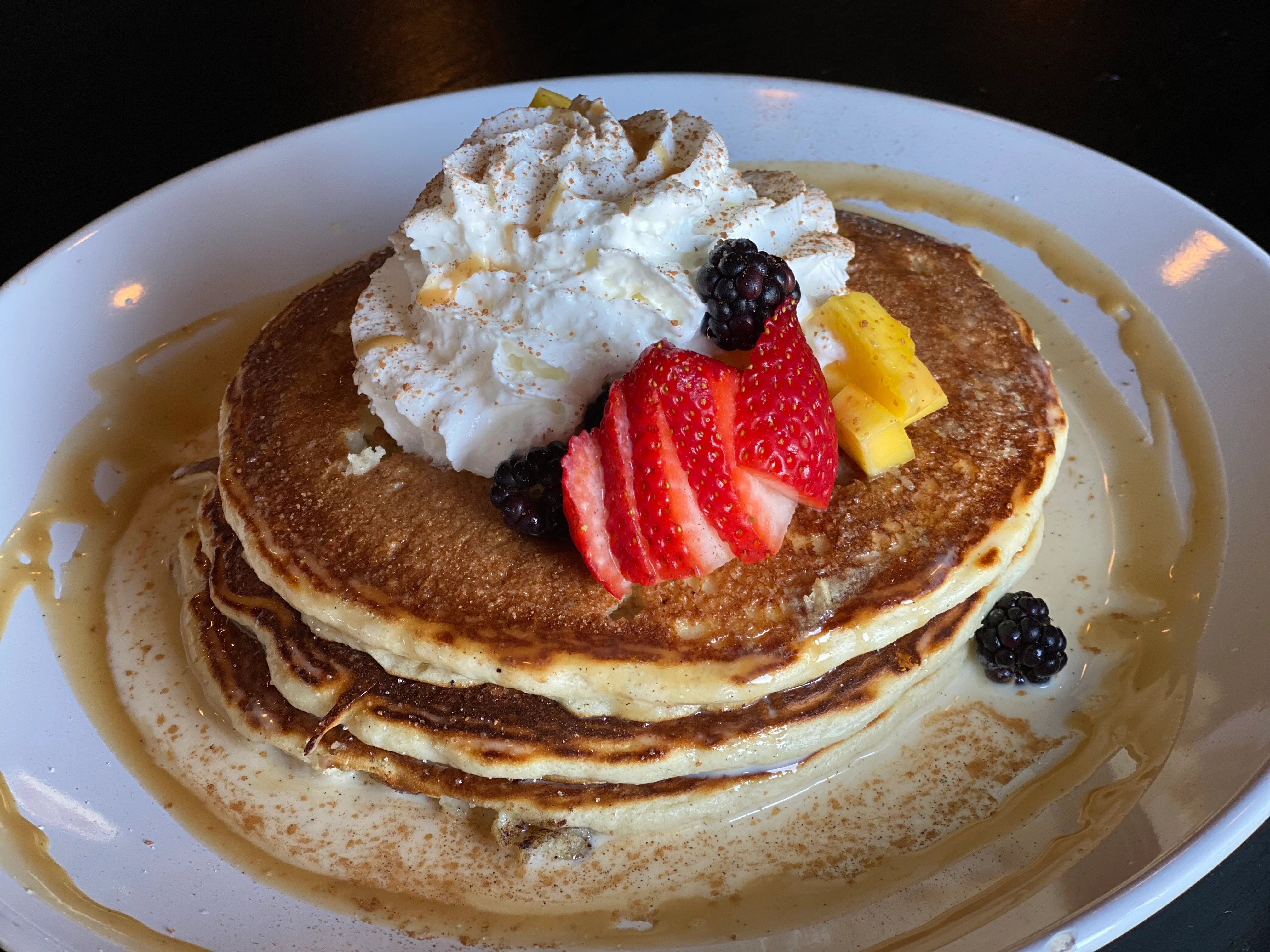 A stack of fluffy pancakes topped with whipped cream and fresh fruit, served with syrup on a white plate.