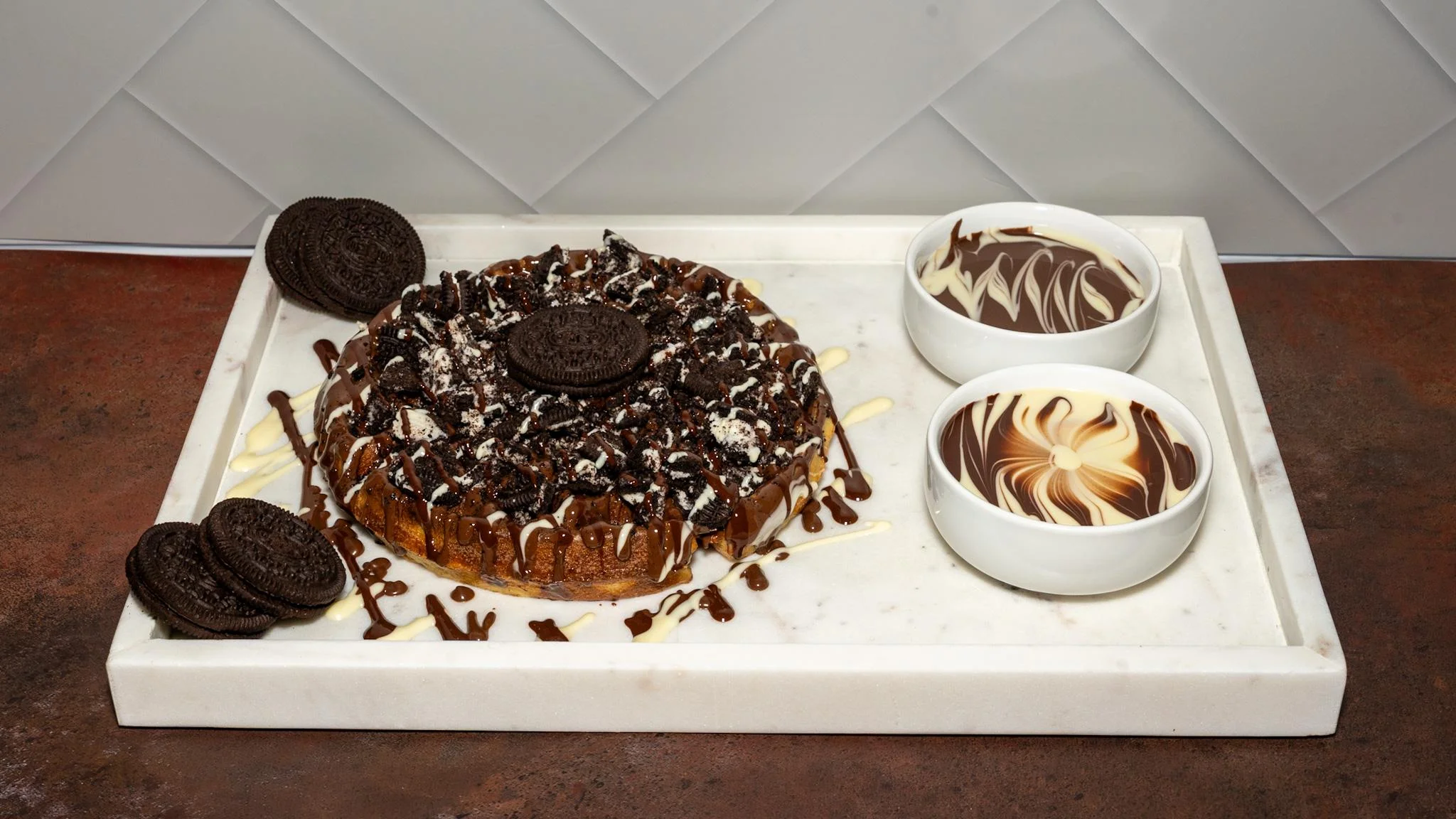 An Oreo dessert pizza topped with crushed Oreos, drizzled with chocolate, and served with two bowls of dipping sauce.