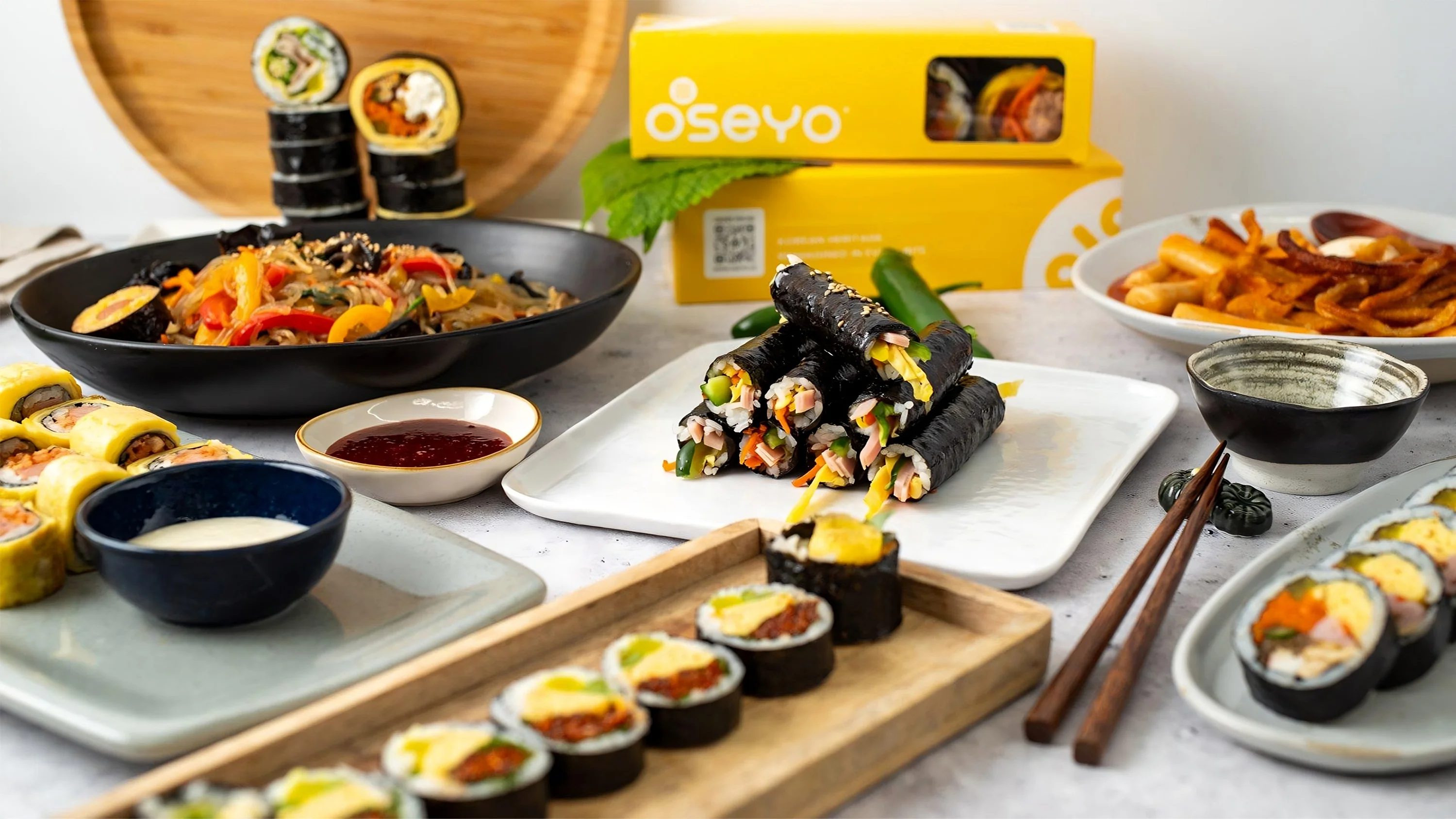 Korean street food spread with kimbap rolls, rice bowls, side dishes, and Oseyo branded takeout boxes.