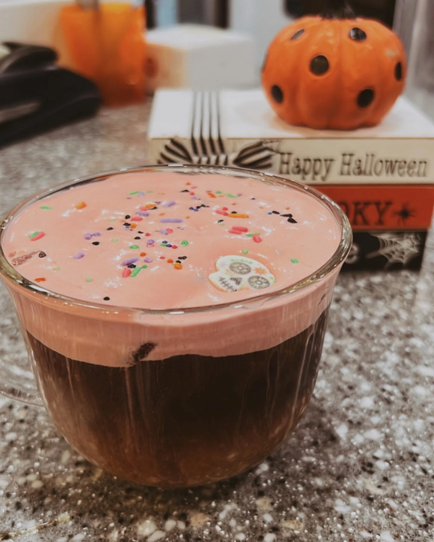 A colorful drink topped with pink froth and sprinkles, served in a glass. In the background, there's a decorative pumpkin and a Halloween-themed display.