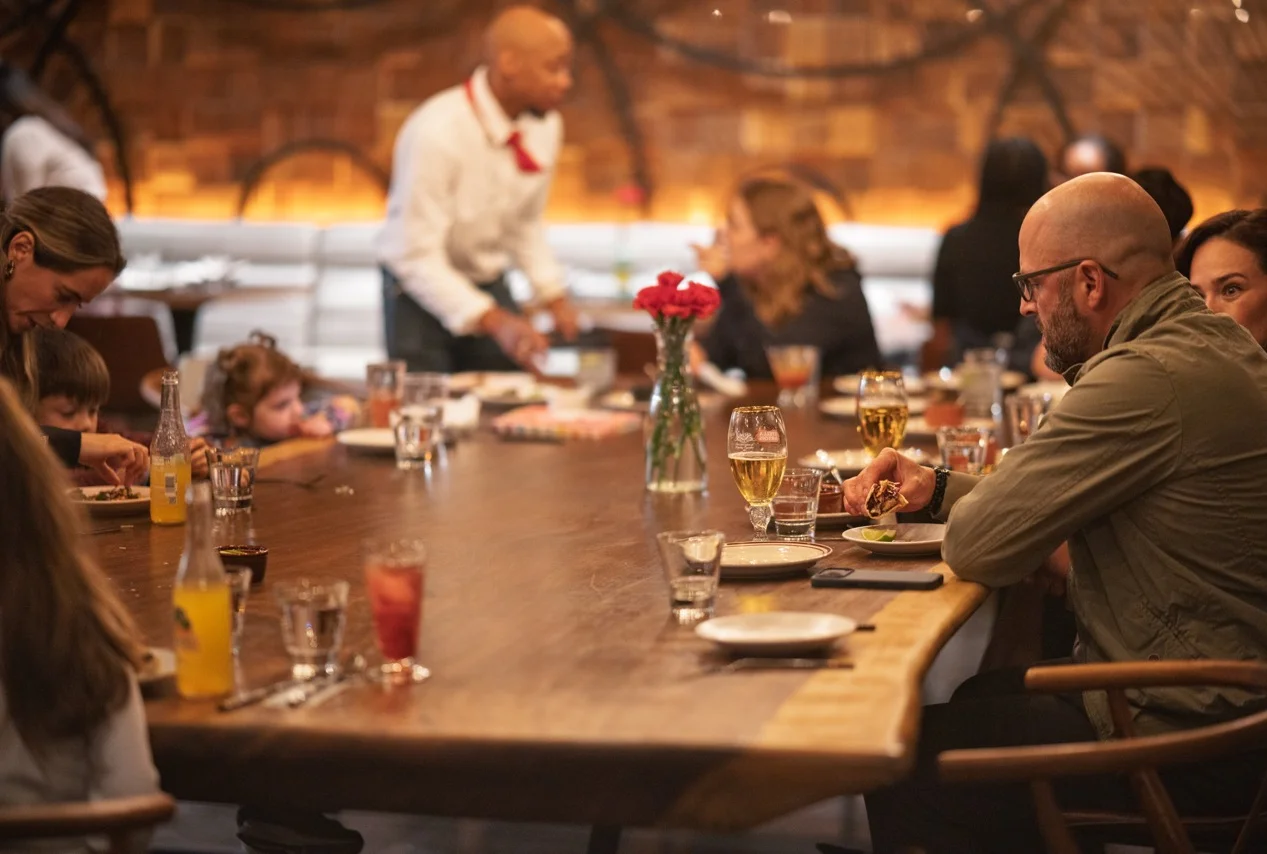 A lively restaurant scene with diners enjoying meals at a long wooden table.