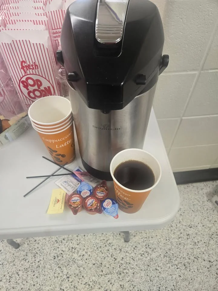 A coffee station featuring a thermal coffee brewer, cups, and creamers on a table. There are prepared cups of coffee and various sweeteners displayed.