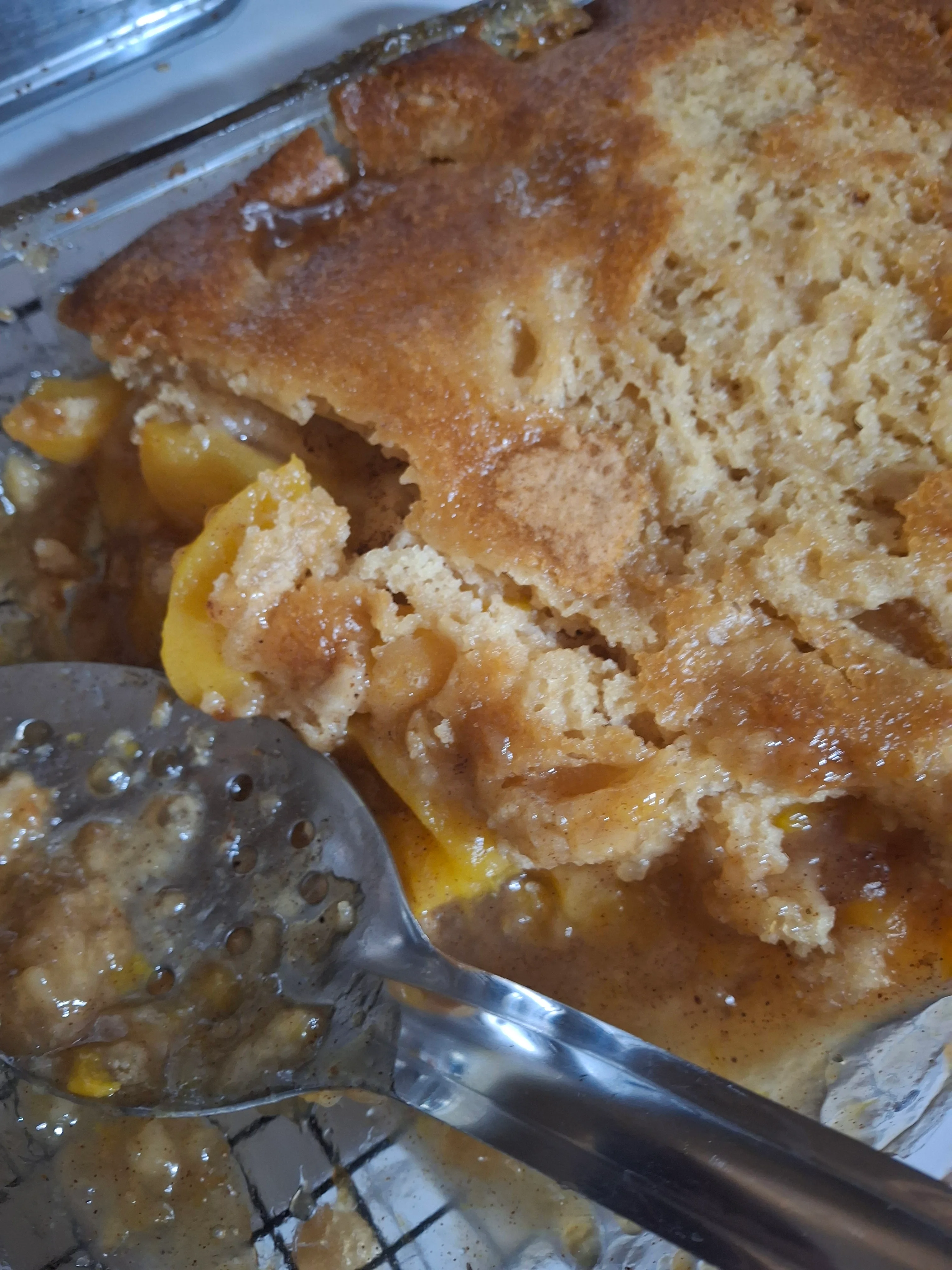 A serving of baked fruit dessert with a golden crust and visible chunks of fruit, served with a slotted spoon.