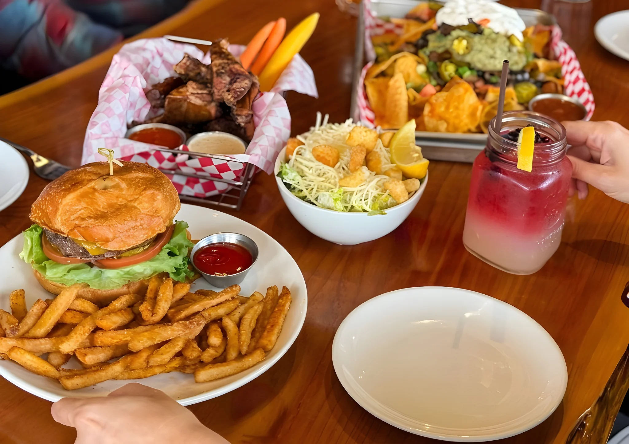 A table with a burger, fries, ribs, nachos, salad, and drinks.