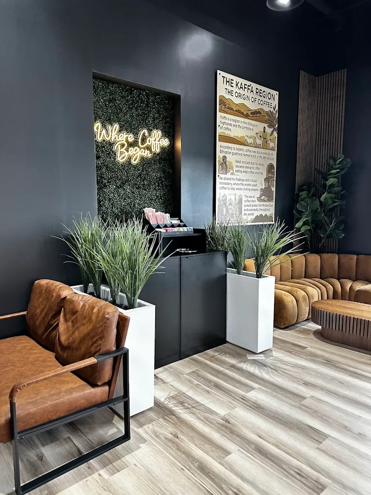 A cozy coffee shop area featuring brown leather seating, plants, and a decorative wall with a neon sign. The interior has a modern aesthetic with wooden flooring.