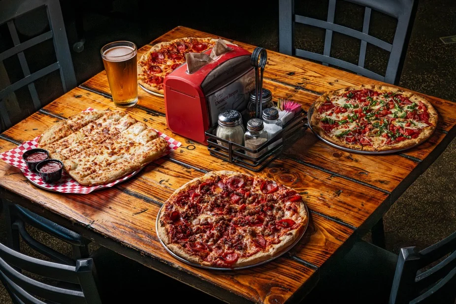 A table with three pizzas, one with pepperoni, one with vegetables, and a cheesy pizza, complemented by a glass of beer and condiments.