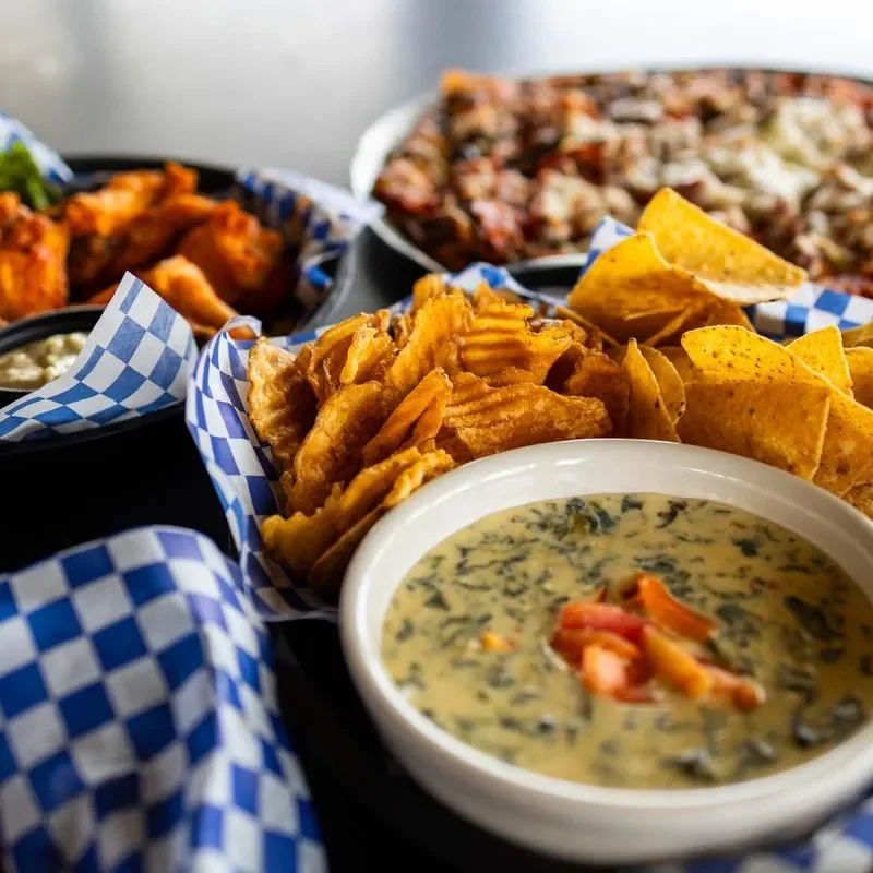 A platter featuring crispy tortilla chips, wavy potato chips, and a creamy spinach dip with diced tomatoes on top. The items are arranged in a vibrant setting.