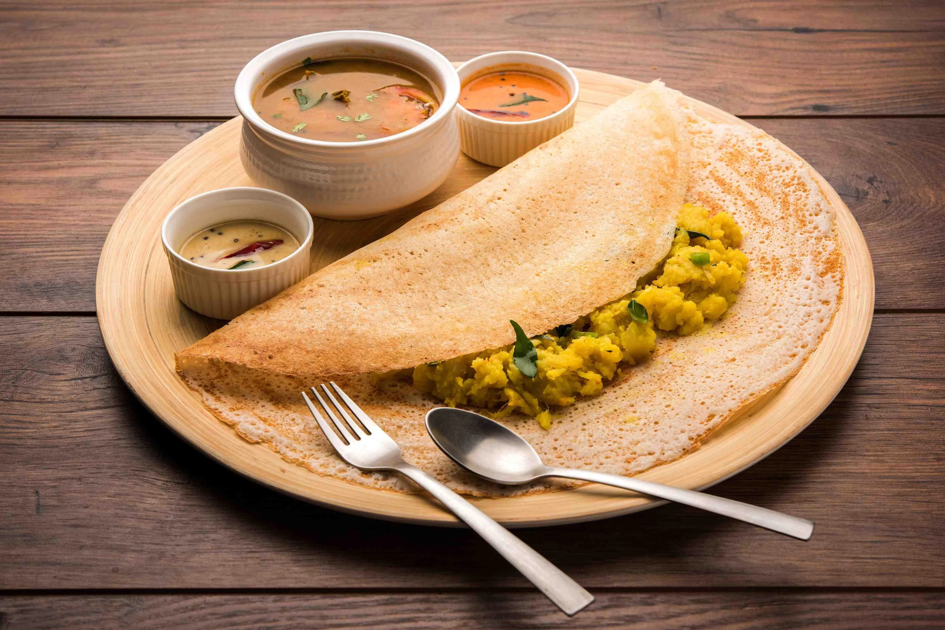 A crispy dosa filled with spiced potatoes, accompanied by sambar and two types of chutney in small bowls.