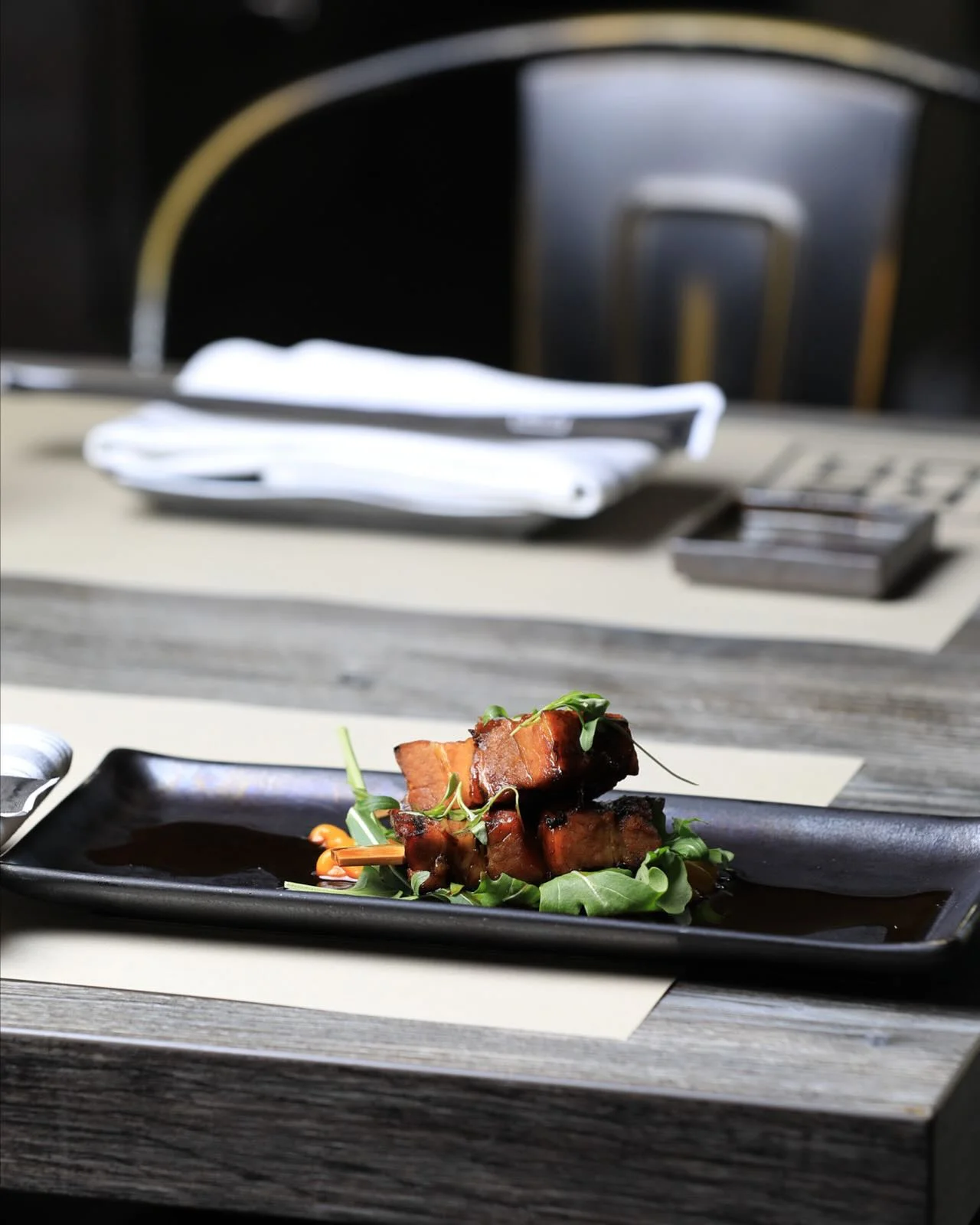 A small stack of glazed, grilled pork belly skewers garnished with microgreens and resting over a bed of fresh arugula on a long black rectangular plate with a dark sauce drizzle, set on a modern restaurant table.