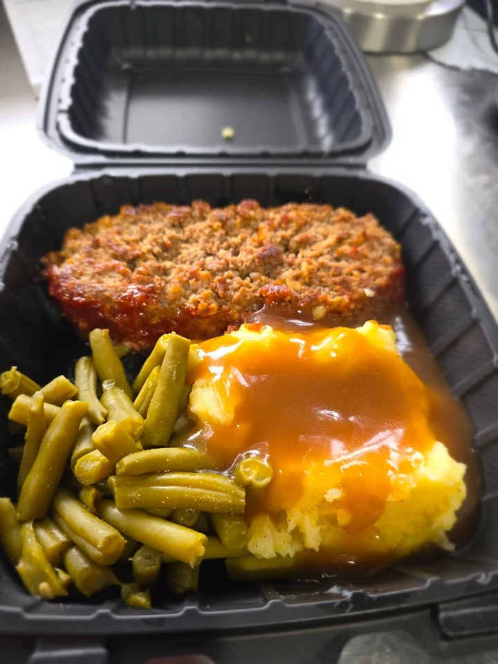 A portion of meatloaf topped with sauce, served with mashed potatoes and green beans in a black takeout container.