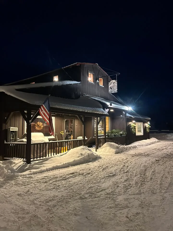 A cozy pub exterior illuminated at night, surrounded by snow. The building features a sign and an American flag.