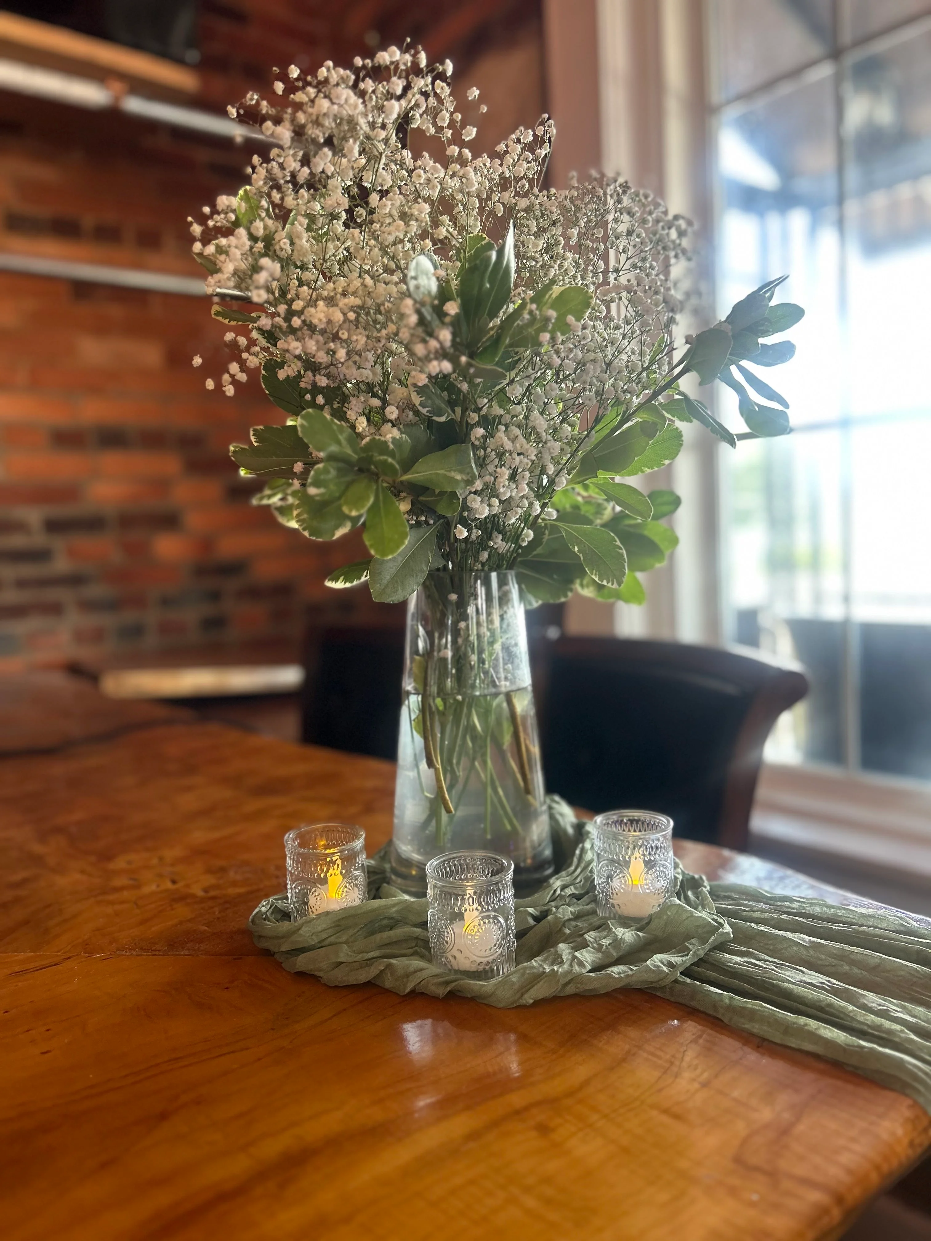 A glass vase filled with delicate white flowers and greenery. The arrangement is set on a wooden table with soft candlelight decorations in the background.