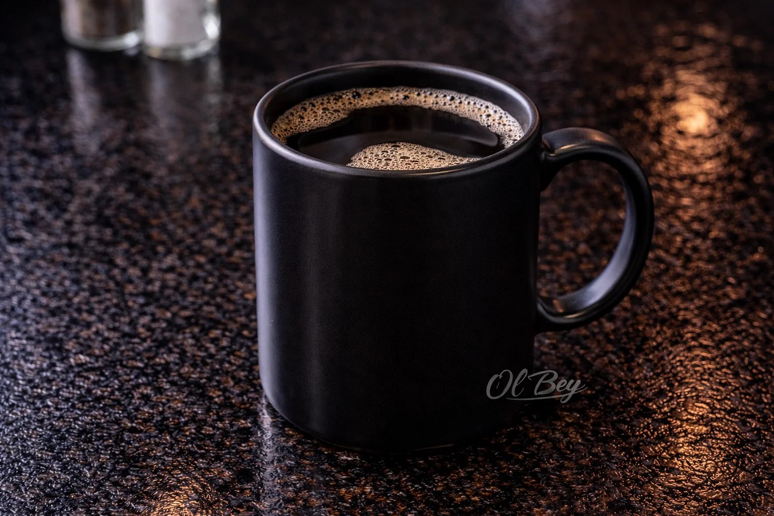A black mug filled with dark coffee, showcasing a frothy surface. The mug is placed on a glossy dark tabletop.