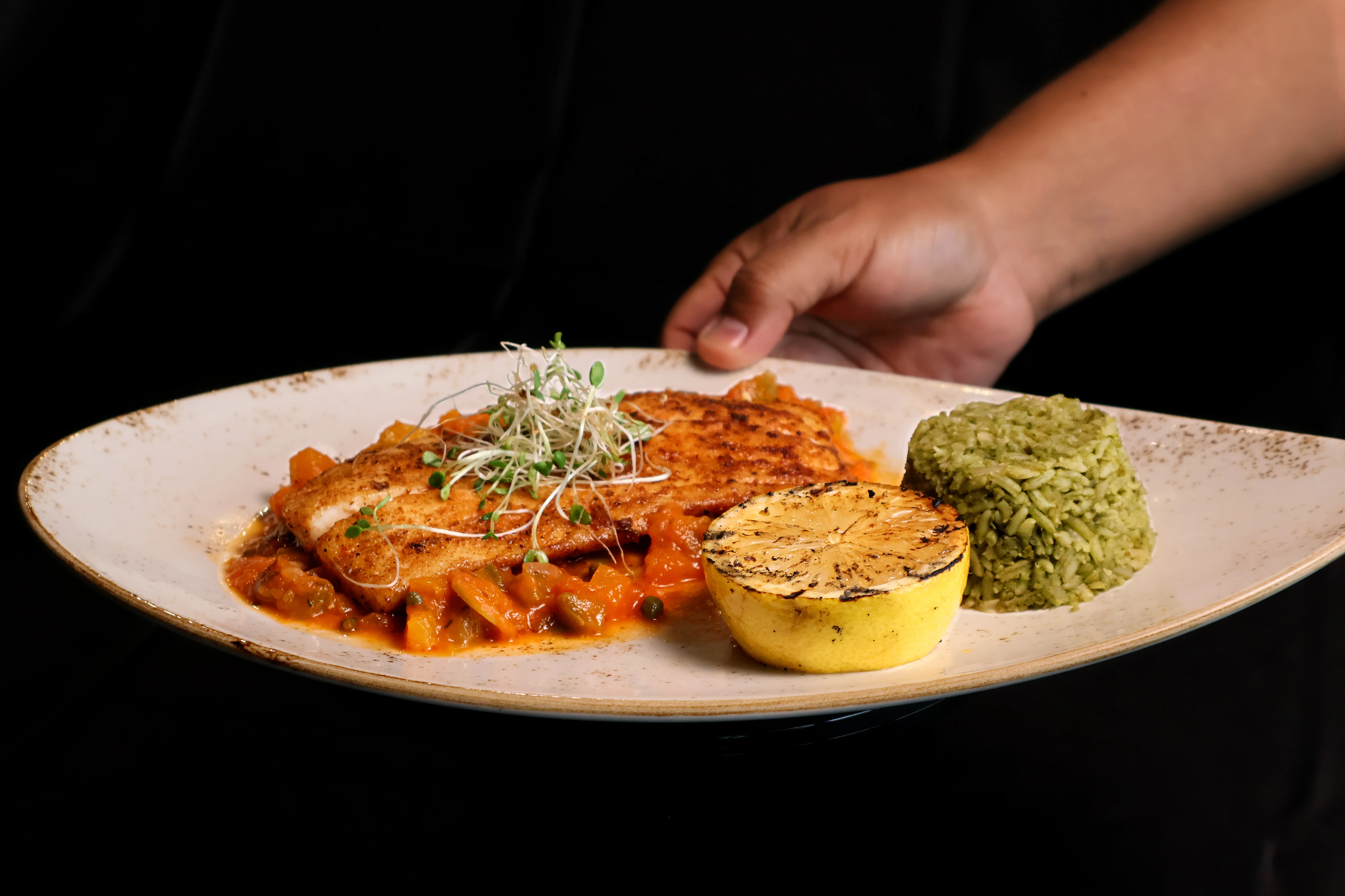 A dish with grilled fish served on a plate with colorful vegetables, a grilled lemon half, and a serving of green rice.