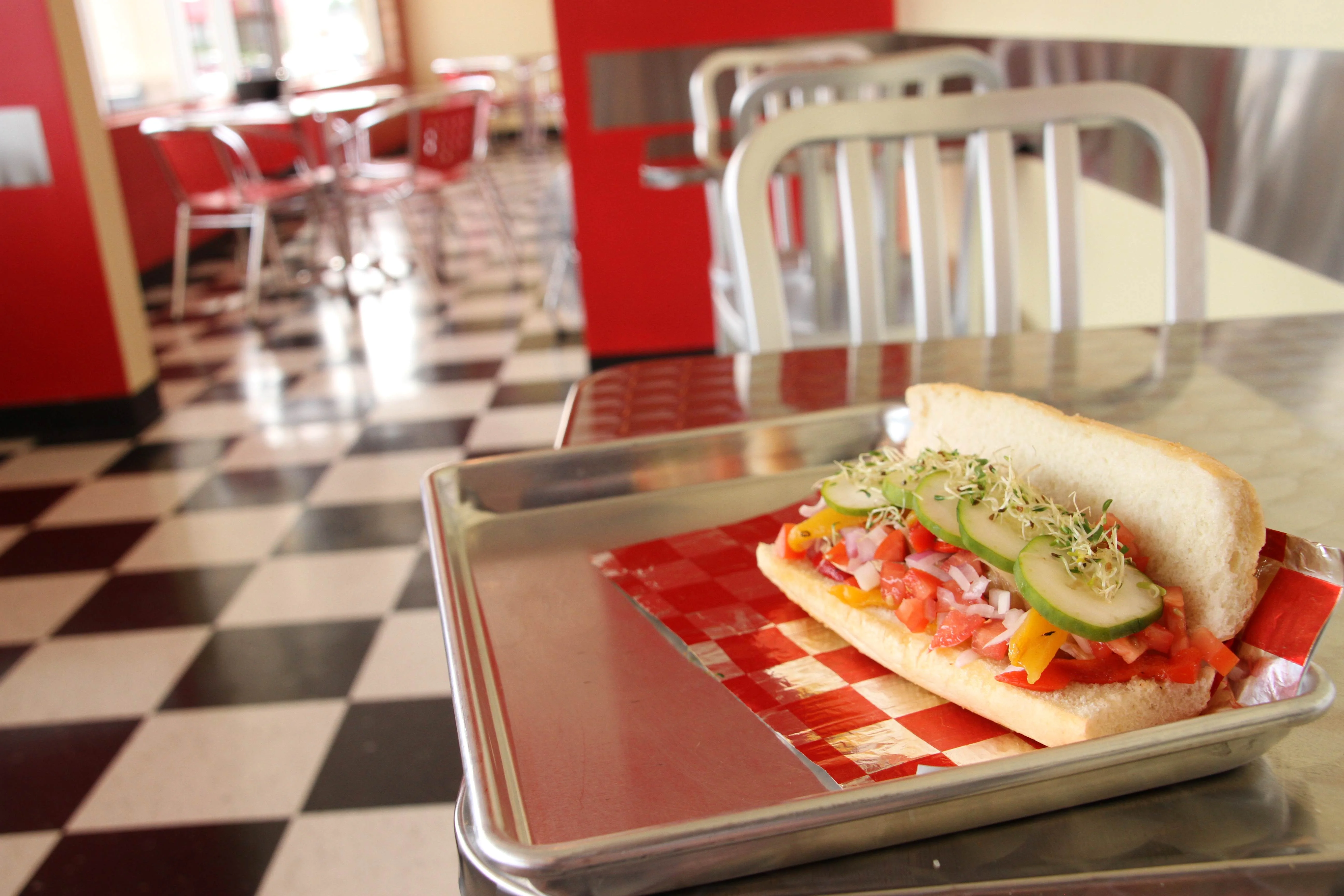 Fresh veggie sub sandwich with diced tomatoes, onions, bell peppers, cucumber slices, and sprouts on a soft hoagie roll, served on a metal tray at TJ’s Sandwich Shop in Northwest Arkansas.