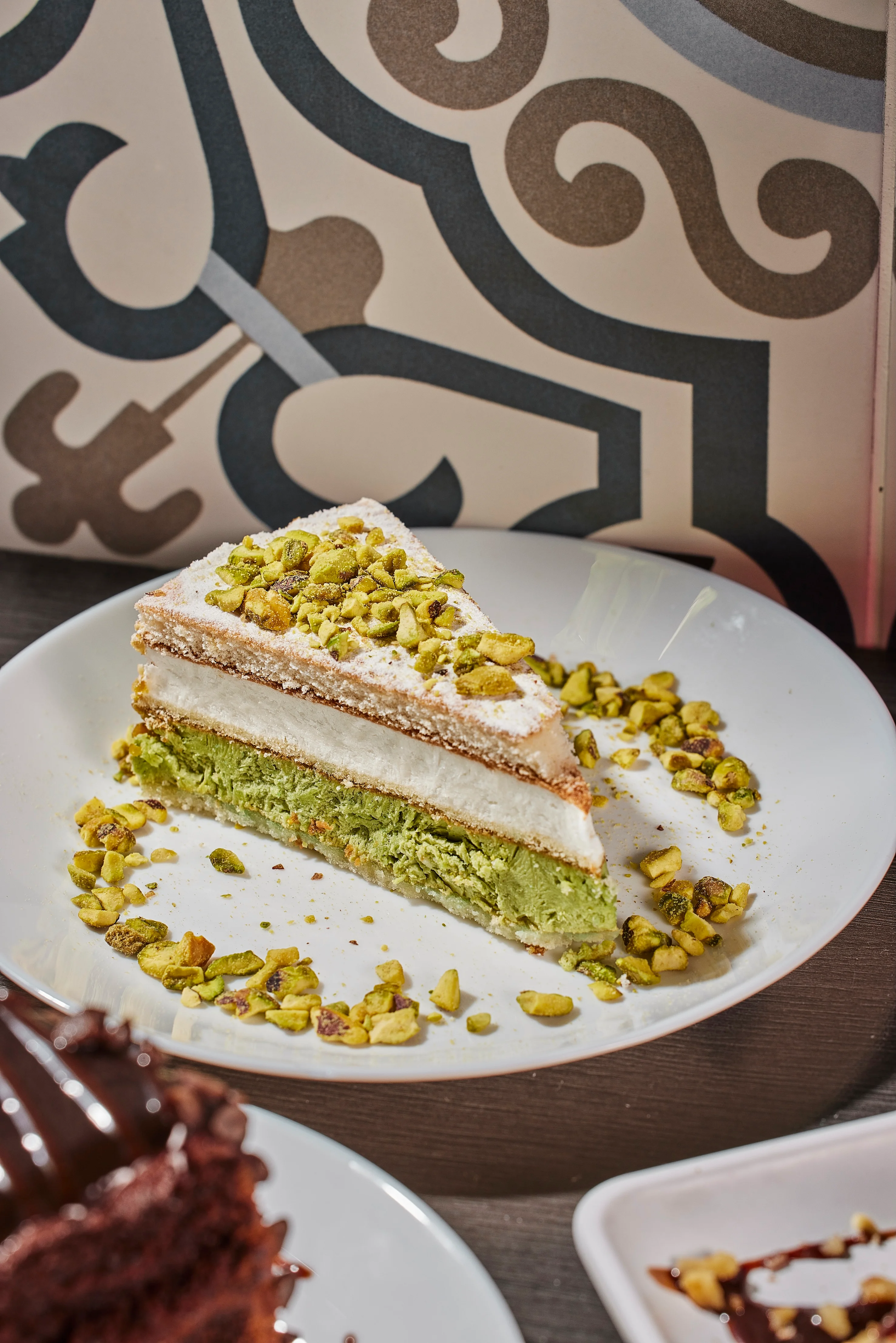 A slice of layered pistachio cake on a white plate, topped with crushed pistachios. Decorative patterned background adds an elegant touch.