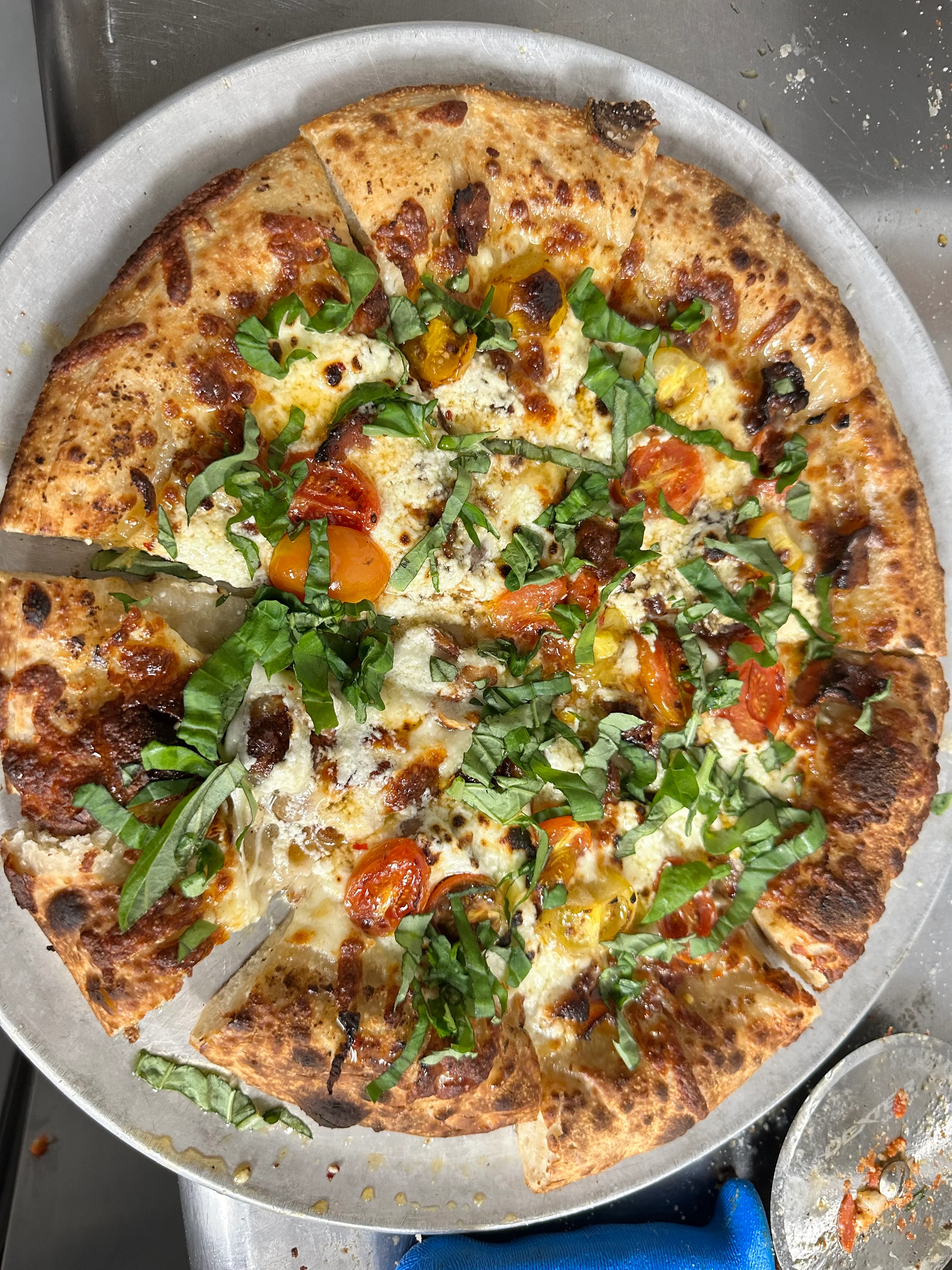 A freshly baked pizza topped with cheese, roasted cherry tomatoes, and fresh greens, sliced and arranged on a metal tray.