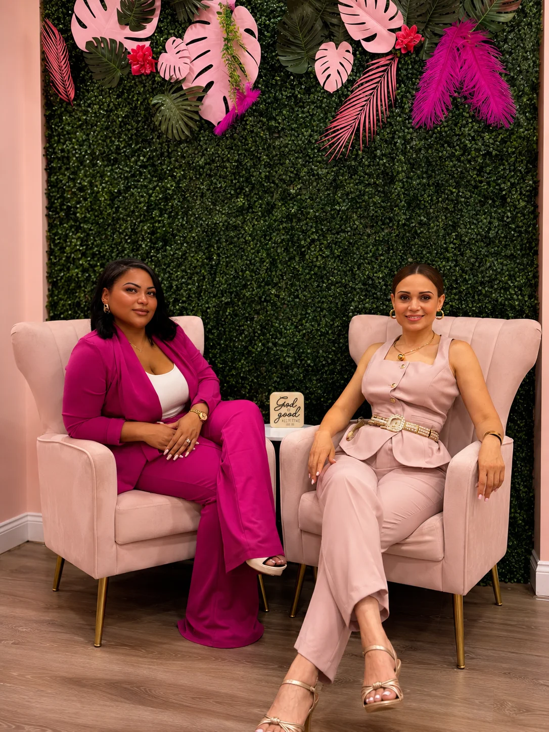 Two women in stylish outfits seated in a chic, floral-decorated space