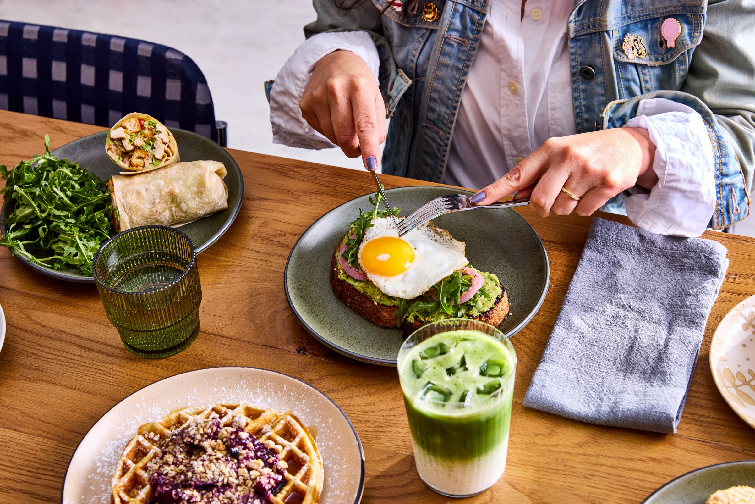 A close-up of a breakfast plate featuring a toasted slice of bread topped with avocado, ham, and a fried egg, along with a side of arugula and a drink.