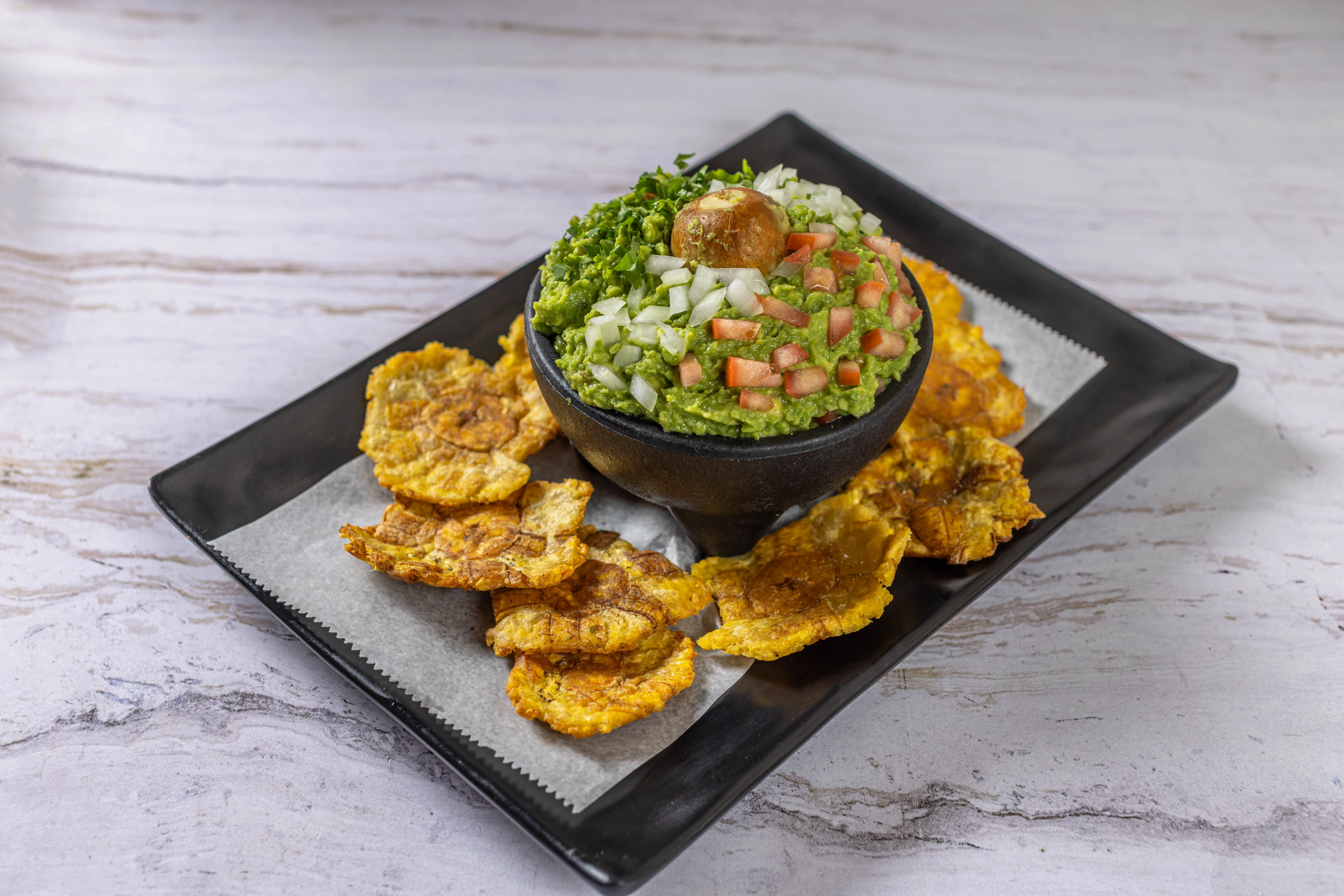 A shareable appetizer of fresh guacamole topped with pico de gallo, served with crispy tostones (fried plantain chips).