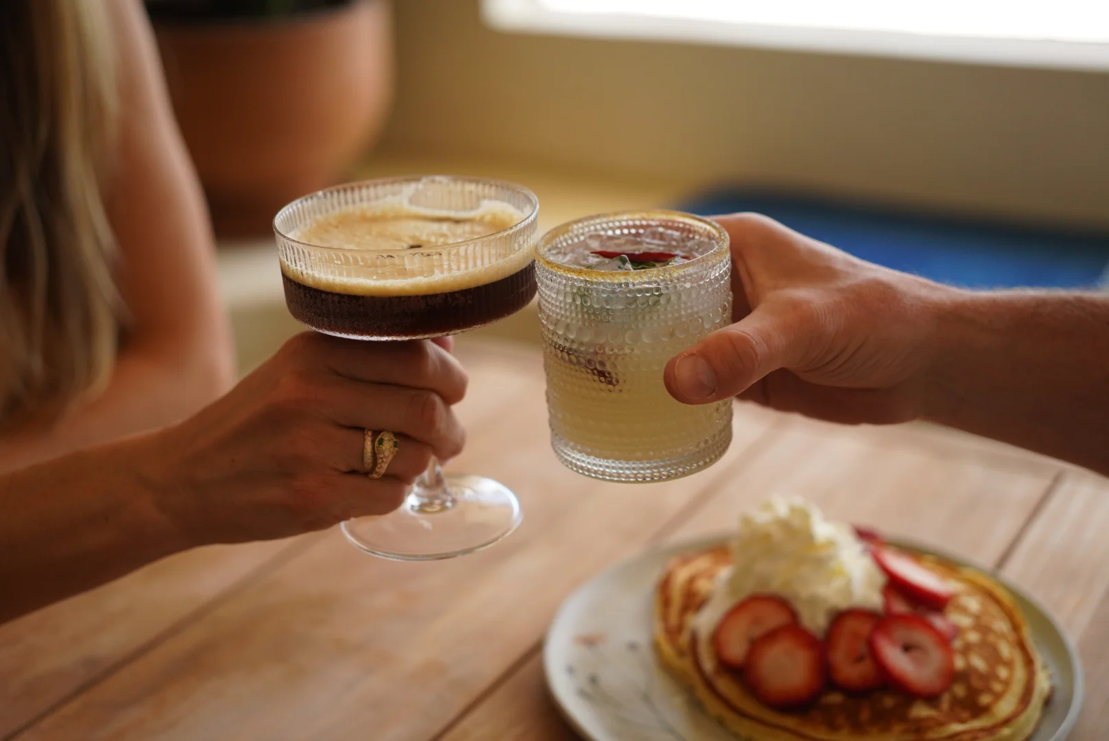 Two cocktails are raised in a toast, one with a frothy top and the other garnished with fruit. A stack of pancakes decorated with strawberries and whipped cream sits nearby.