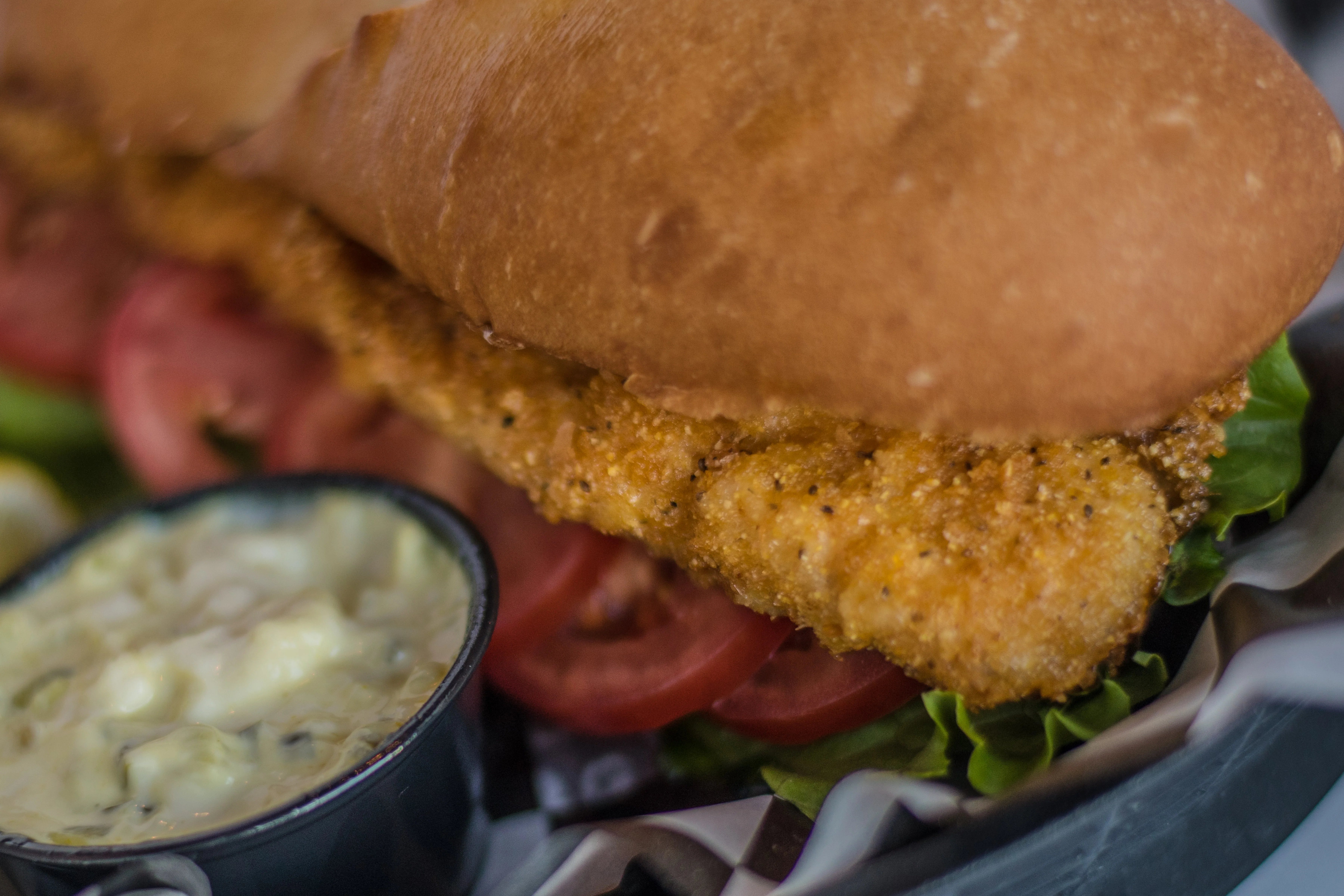 A sandwich filled with a breaded fish fillet, lettuce, and tomato, accompanied by a dipping sauce.