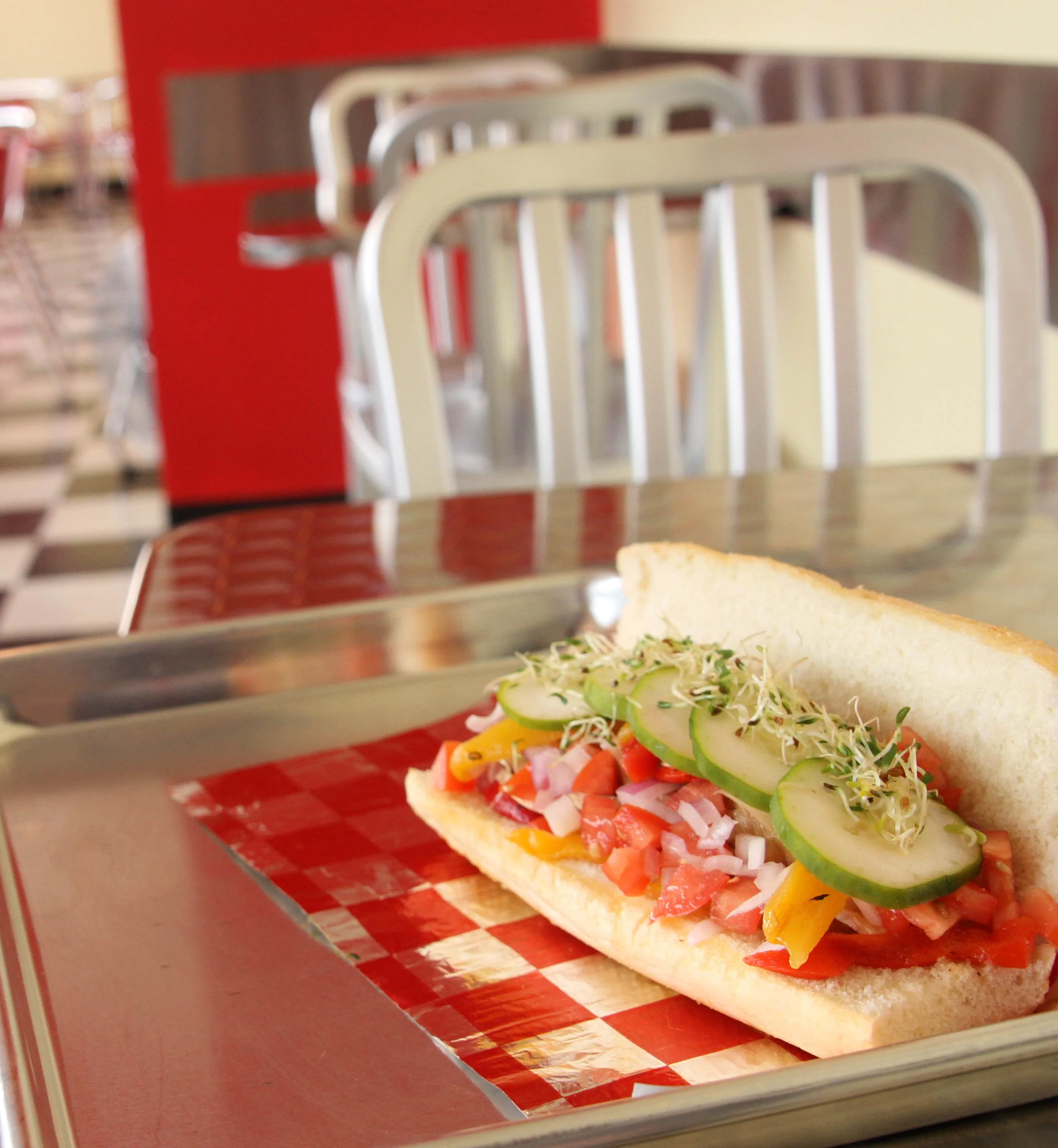 Fresh vegetable sub sandwich with cucumbers, sprouts, diced tomatoes, onions, and bell peppers on a hoagie roll served on a metal tray in a retro-style diner with checkered floors and aluminum chairs.