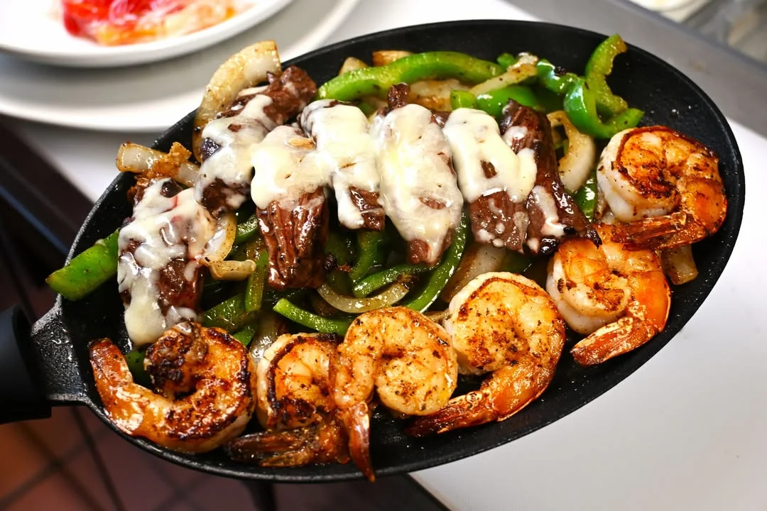 A sizzling platter of shrimp and beef, topped with melted cheese, accompanied by sautéed onions and green peppers.