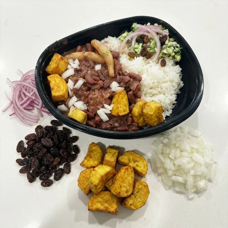 A bowl with rice, red beans, pieces of plantain, and garnish of onions and raisins, presented alongside additional ingredients.