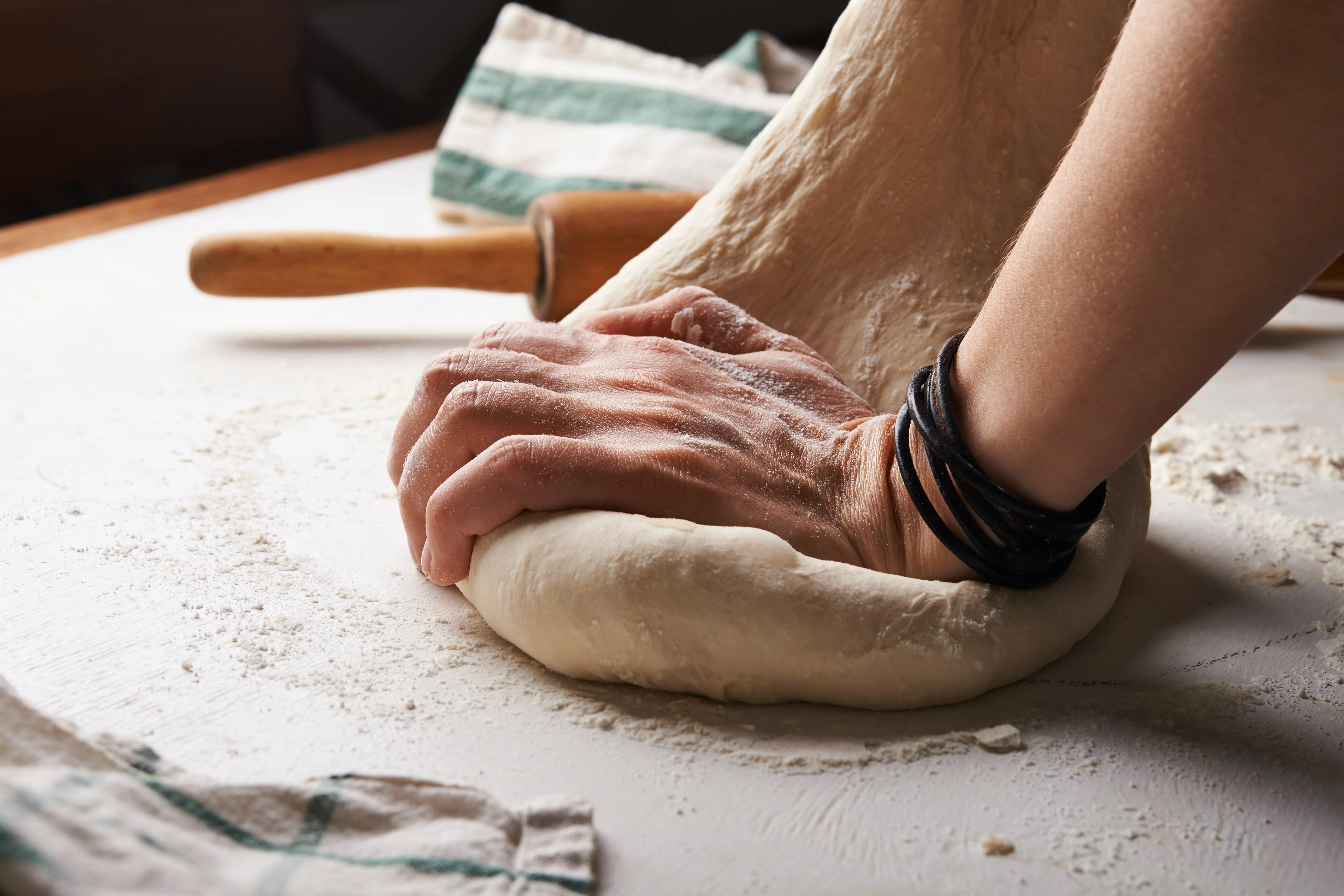 A close-up of a hand kneading dough on a floured surface, with a rolling pin in the background.