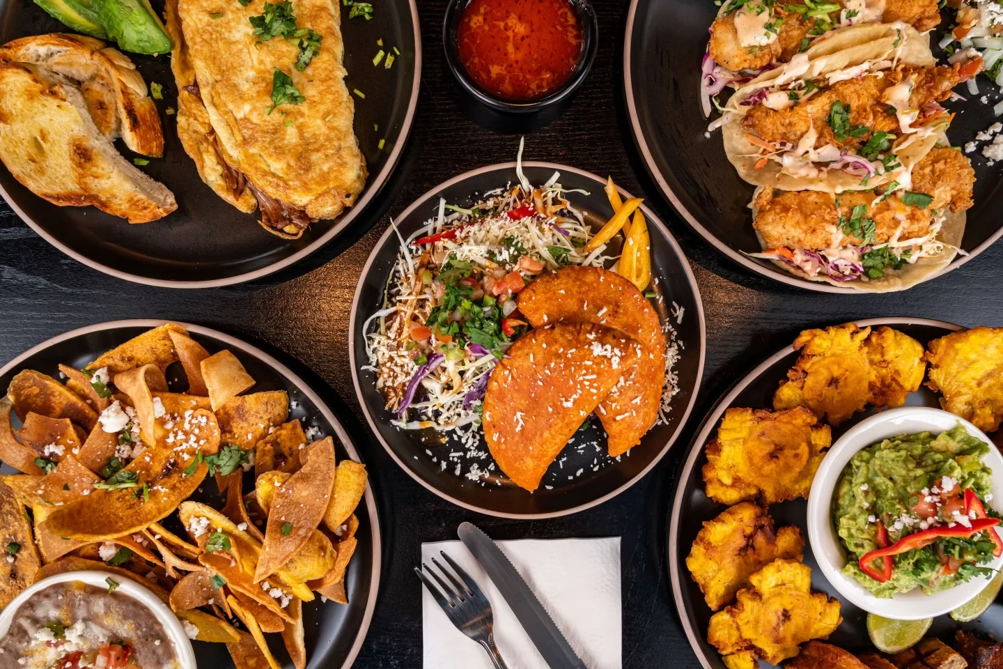 A vibrant spread of Mexican cuisine featuring tacos, tostadas, tostones, and guacamole. Each dish is beautifully arranged on black plates with fresh toppings.