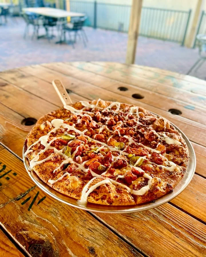 A pizza topped with a variety of meats and drizzled with white sauce, served on a metallic tray outdoors.