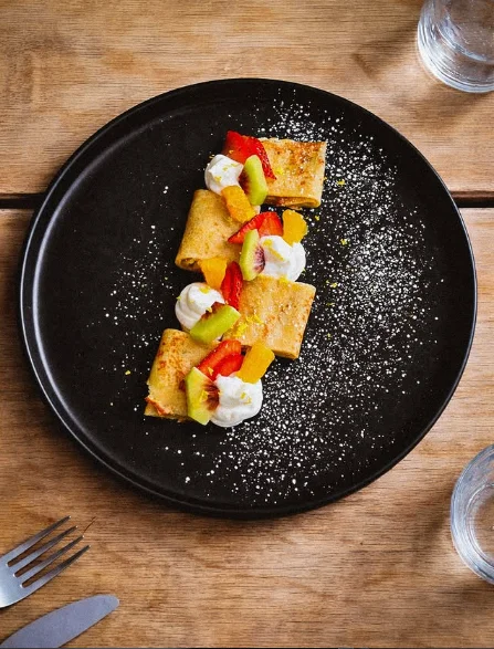 A plate of crepes topped with a mix of colorful fruits and whipped cream, elegantly presented on a black plate.