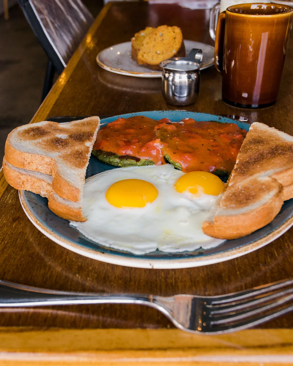 A breakfast plate featuring two sunny-side-up eggs, crispy toast, and a savory dish topped with tomato sauce and served with green ingredients.