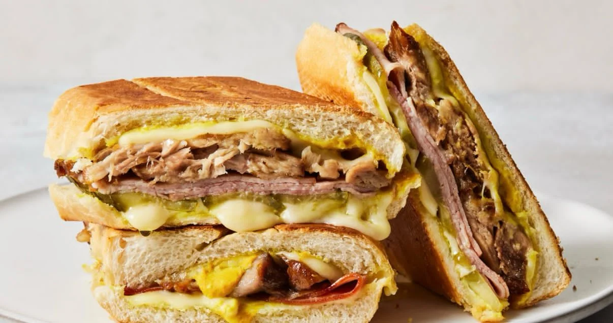A close-up view of a Cuban sandwich cut in half, showcasing layers of roasted pork, ham, melted cheese, pickles, and mustard, pressed between toasted bread with a golden-brown crust.