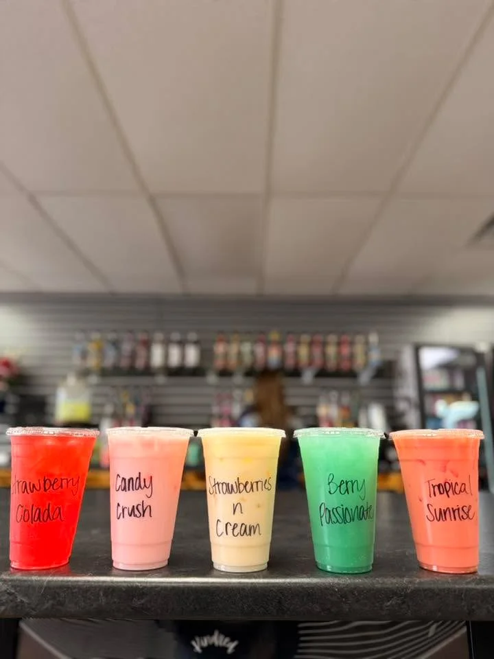 Five colorful drinks lined up on a counter, each in clear cups with handwritten labels, showcasing various fruity flavors.