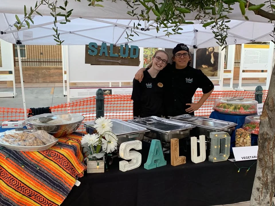 A vibrant food booth named 'Salud' featuring two smiling staff members. The booth is decorated with colorful textiles and showcases various food trays under a white canopy.