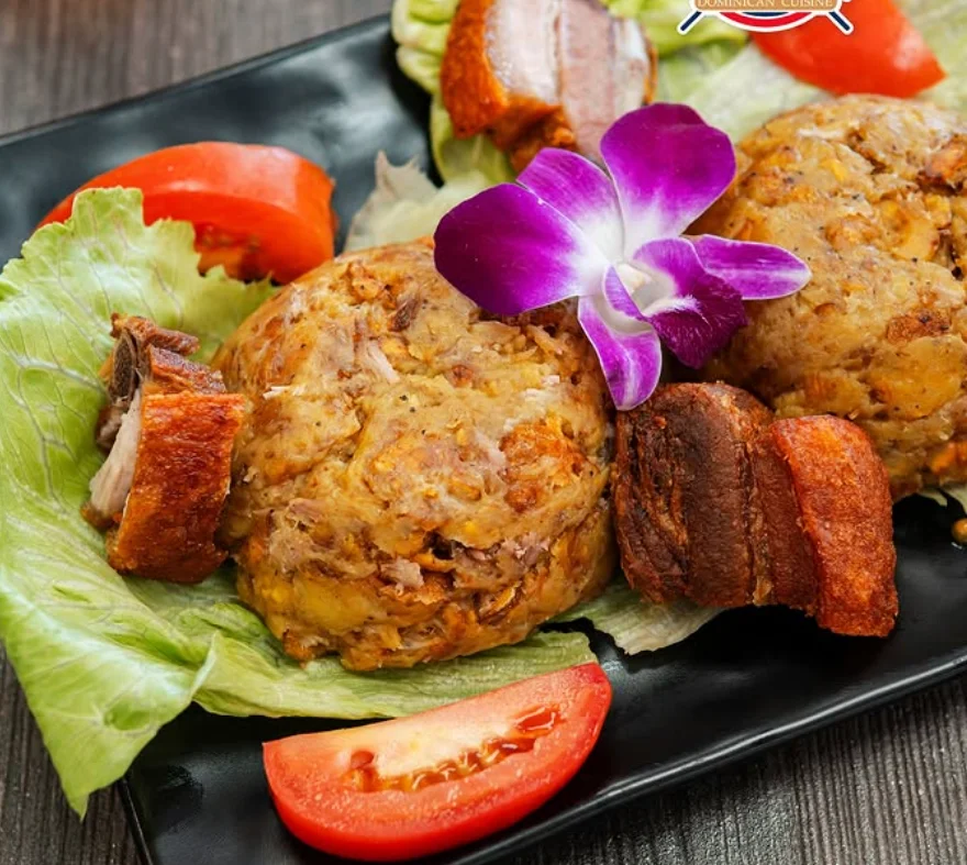 A dish featuring two servings of mofongo, garnished with lettuce, tomato slices, and a purple flower. Crispy pork pieces accompany the mofongo on a black plate.