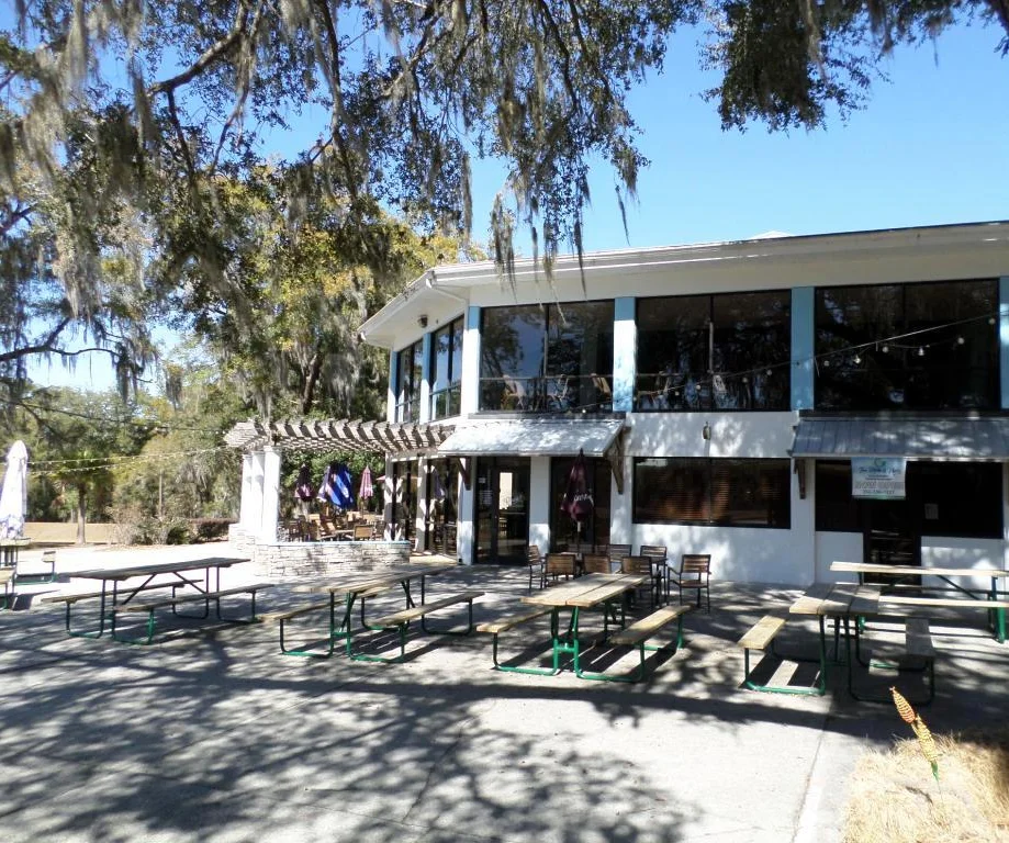 An outdoor dining area featuring tables and umbrellas in front of a restaurant with large windows. The establishment has a welcoming sign and a spacious patio.