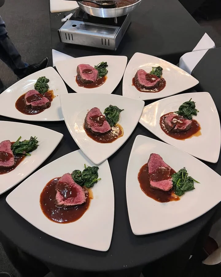 Eight white triangular plates are arranged in a circle on a black tablecloth, each featuring a gourmet serving of sliced rare beef topped with a rich brown sauce and a side of sautéed greens. A stainless steel pan sits on a burner in the background, indicating the dish was freshly prepared for a tasting or upscale event.